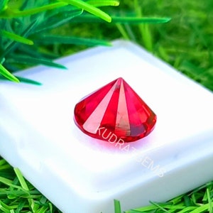 May include: A faceted, round-cut vibrant red gemstone with a pointed crown, resting on a white square display base. The gemstone features the text KUDRATGEMS etched across its surface. The object is displayed against a bright green background.