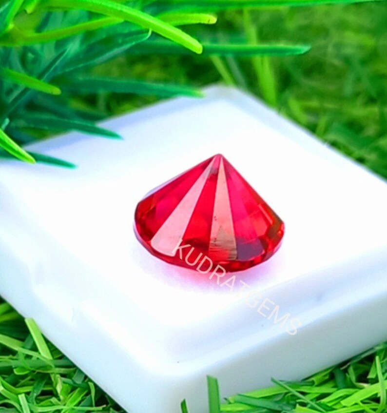 May include: A faceted, round-cut vibrant red gemstone with a pointed crown, resting on a white square display base. The gemstone features the text KUDRATGEMS etched across its surface. The object is displayed against a bright green background.