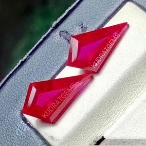 Puede incluir: Dos piedras preciosas rojas en forma de cometa con superficies facetadas. Cada piedra tiene el texto KUDRATGEMS impreso en blanco en la parte superior. Las gemas se muestran sobre una superficie blanca texturizada con un borde azul oscuro.