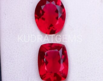 May include: Two vibrant red gemstones with faceted surfaces. The top stone is oval-shaped, while the bottom stone features a cushion cut. The text KUDRATGEMS is visible in white lettering across the image, overlaying the two loose red gems.