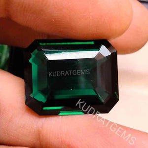 May include: A faceted, emerald-cut gemstone with a deep green hue. The gem is held between two fingers, showcasing its rectangular shape and internal facets. The text "KUDRAT GEMS" is visible on the gem's surface.