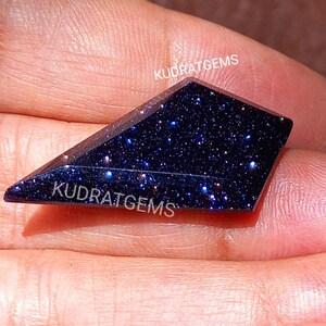 May include: A kite-shaped blue sandstone gemstone with a dark, sparkling surface featuring numerous reflective glitter-like inclusions. The stone is faceted with sharp edges and displays the white text KUDRATGEMS printed on its surface.