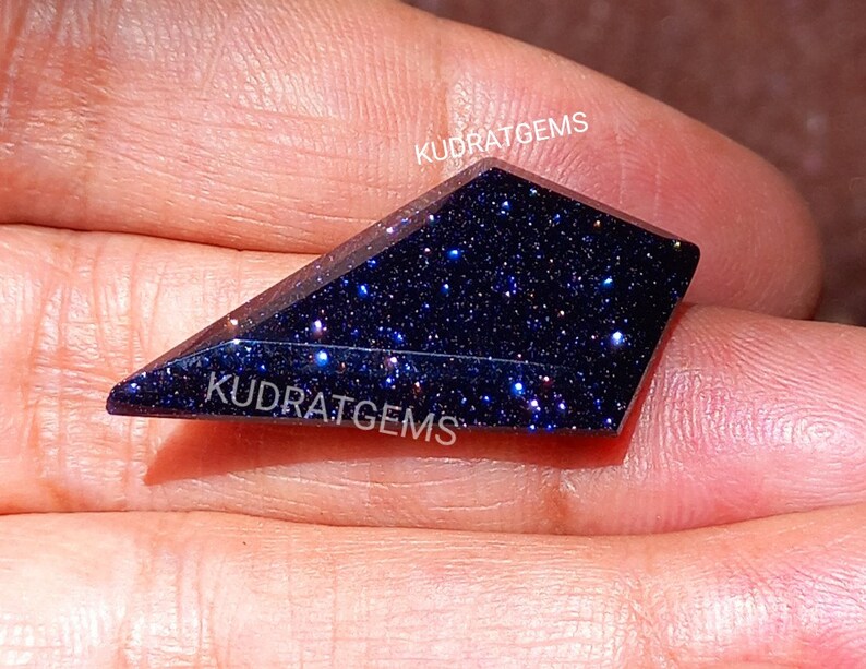 May include: A kite-shaped blue sandstone gemstone with a dark, sparkling surface featuring numerous reflective glitter-like inclusions. The stone is faceted with sharp edges and displays the white text KUDRATGEMS printed on its surface.