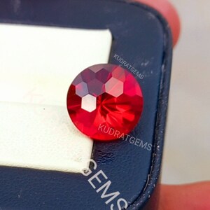 May include: A round, faceted, vibrant red gemstone with a brilliant cut that reflects light across its surface. The gemstone is positioned on a white and dark blue display case, which features the text KUDRATGEMS printed in white lettering.