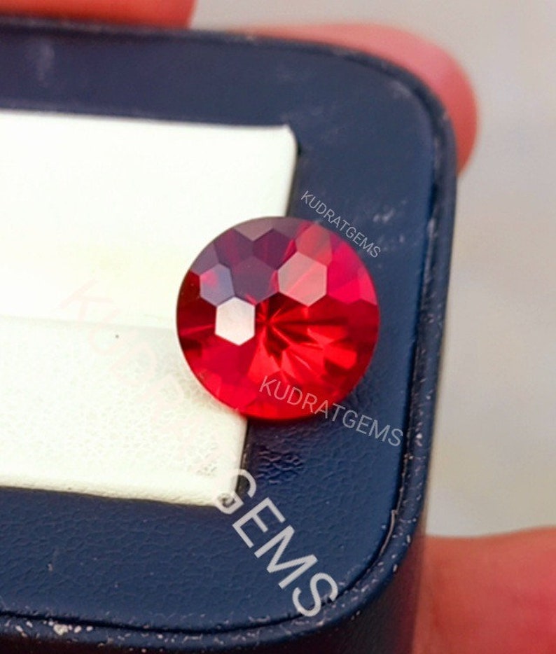 May include: A round, faceted, vibrant red gemstone with a brilliant cut that reflects light across its surface. The gemstone is positioned on a white and dark blue display case, which features the text KUDRATGEMS printed in white lettering.