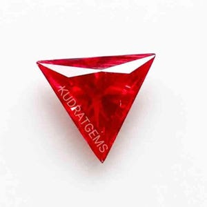 May include: A vibrant, triangular-cut red gemstone with a faceted surface that reflects light. The word KUDRATGEMS is printed in white text diagonally across the center of the stone, which features sharp, pointed corners and a deep crimson hue.
