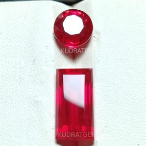 Könnte beinhalten: Zwei rote Edelsteine, einer rund und einer rechteckig, werden vor einem weißen Hintergrund gezeigt. Der runde Edelstein hat eine facettierte Oberfläche und der Text "KUDRATO" ist sichtbar. Der rechteckige Edelstein hat eine glatte Oberfläche und der Text "KUDRATGEN" ist sichtbar.