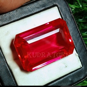 May include: A vibrant, rectangular-cut, red gemstone with a polished surface. The gem is displayed in a black case with a white interior. The text "KUDRATGEM" is visible on the gem's surface.