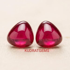 May include: Two polished, triangular-shaped, deep red gemstones. The gems have a smooth, glossy surface and are set against a neutral background. The text "KUDRAT GEMS" is visible at the bottom.