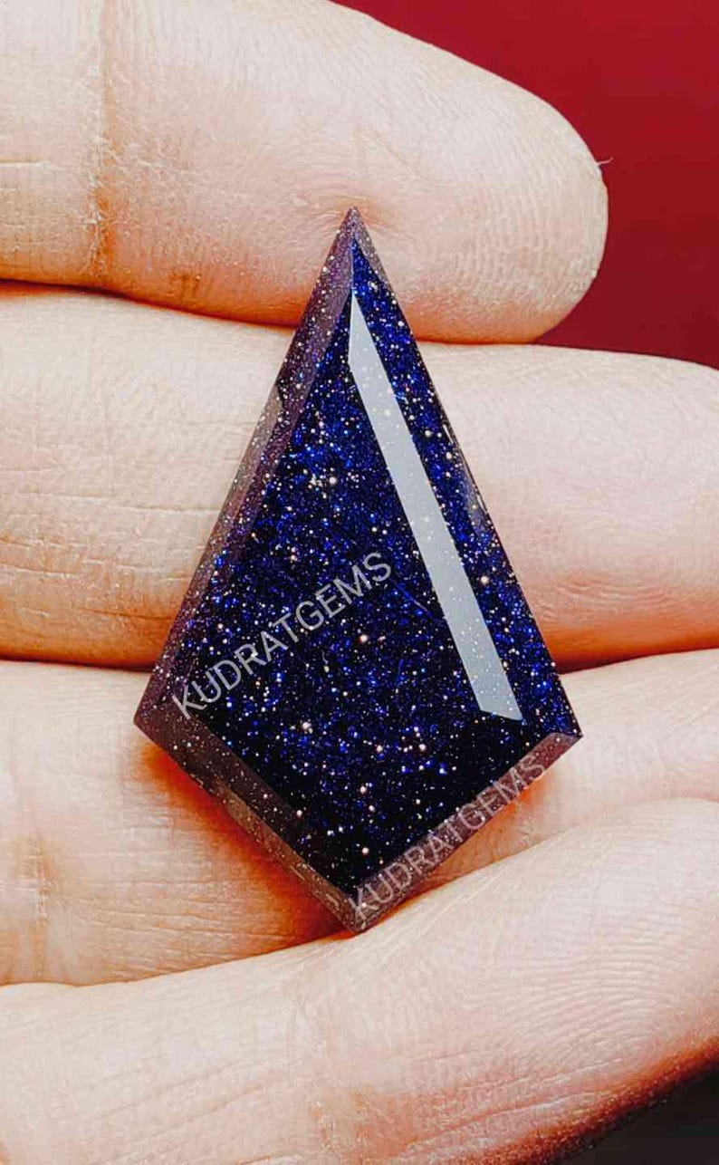 May include: A kite-shaped blue sandstone gemstone with a dark blue base and sparkling metallic flecks throughout. The surface features a faceted cut and the text KUDRATGEMS is printed twice across the face of the stone in white lettering.