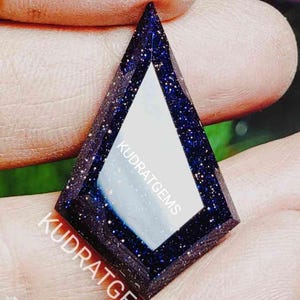 May include: A kite-shaped blue sandstone gemstone with a smooth, reflective center and sparkling metallic inclusions throughout the dark blue surface. The text KUDRATGEMS is printed in white across the face of the stone.