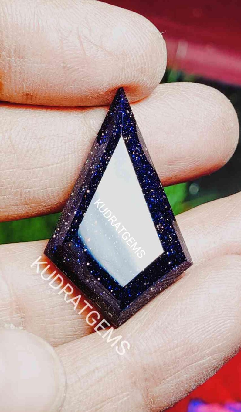 May include: A kite-shaped blue sandstone gemstone with a smooth, reflective center and sparkling metallic inclusions throughout the dark blue surface. The text KUDRATGEMS is printed in white across the face of the stone.
