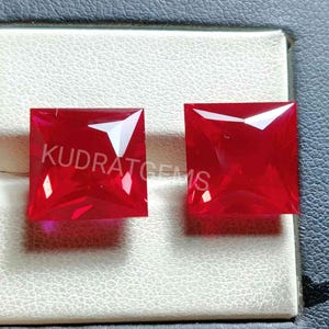 May include: Two square-shaped, red gemstones with a faceted cut. The gemstones are displayed on a white surface.