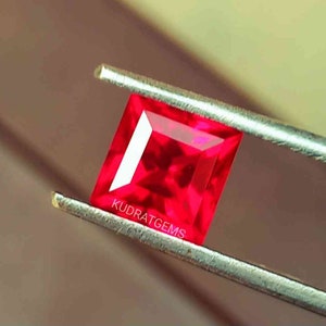 May include: A vibrant, rectangular-cut ruby gemstone held by tweezers. The gem is a deep, saturated red with a bright, reflective surface. The text "KUDRATGEMS" is visible on the gem. The gemstone is a jewelry supply.