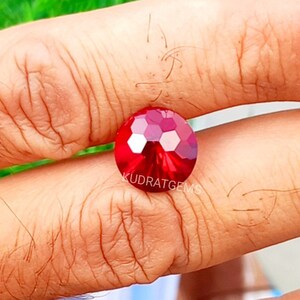 May include: A round, faceted, vibrant red gemstone featuring a brilliant cut with multiple reflective surfaces. The stone is marked with the text KUDRATGEMS printed in white across its center, showcasing a deep crimson hue and high clarity.