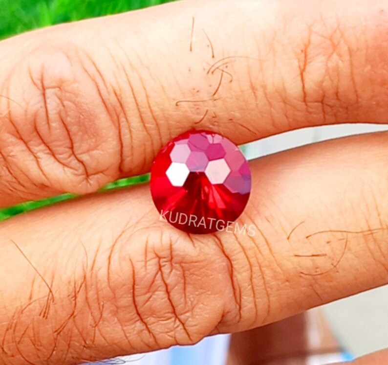 May include: A round, faceted, vibrant red gemstone featuring a brilliant cut with multiple reflective surfaces. The stone is marked with the text KUDRATGEMS printed in white across its center, showcasing a deep crimson hue and high clarity.