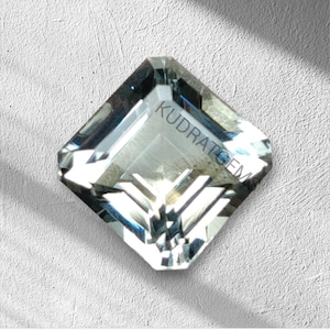 Könnte beinhalten: Ein facettierter, quadratisch geschliffener Edelstein mit einem blassgrünen Farbton. Der Stein hat eine klare, reflektierende Oberfläche mit dem Text "KUDRATGEM" auf der Oberseite. Der Edelstein liegt vor einem strukturierten, weißen Hintergrund.