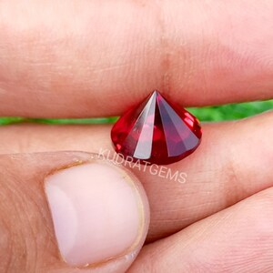 May include: A faceted, round-cut, deep red gemstone with a brilliant, reflective surface. The stone features a pointed pavilion and a flat table, displaying intense crimson hues. The text KUDRATGEMS is printed in white across the center of the gem.