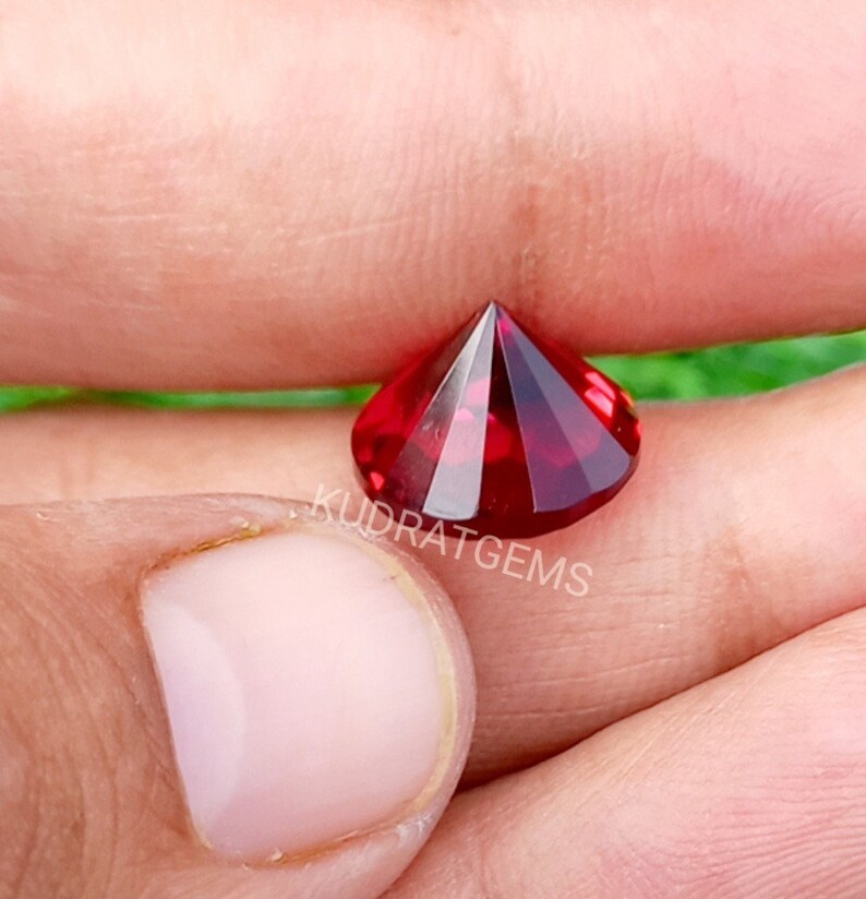 May include: A faceted, round-cut, deep red gemstone with a brilliant, reflective surface. The stone features a pointed pavilion and a flat table, displaying intense crimson hues. The text KUDRATGEMS is printed in white across the center of the gem.