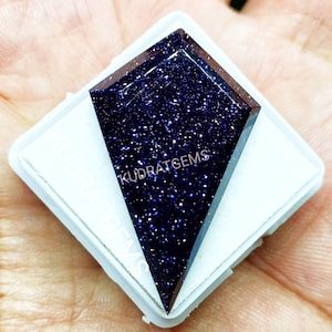 May include: A kite-shaped blue sandstone gemstone with a sparkling, glittery surface texture. The dark blue stone is set against a white square display card featuring the text KUDRATGEMS printed in white lettering across the surface.