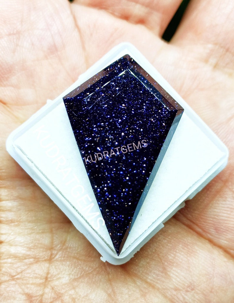May include: A kite-shaped blue sandstone gemstone with a sparkling, glittery surface texture. The dark blue stone is set against a white square display card featuring the text KUDRATGEMS printed in white lettering across the surface.