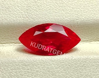 May include: A marquise-cut, vibrant red gemstone featuring a faceted surface that reflects light. The word KUDRATGEM is printed in white letters across the center of the stone, which has a pointed, elongated shape with symmetrical sides.