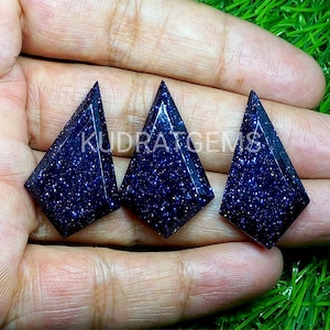 May include: Three kite-shaped blue goldstone cabochons with a deep navy color and sparkling metallic inclusions. Each gemstone features a faceted surface and a pointed top. The text KUDRATGEMS is overlaid across the center of the stones.