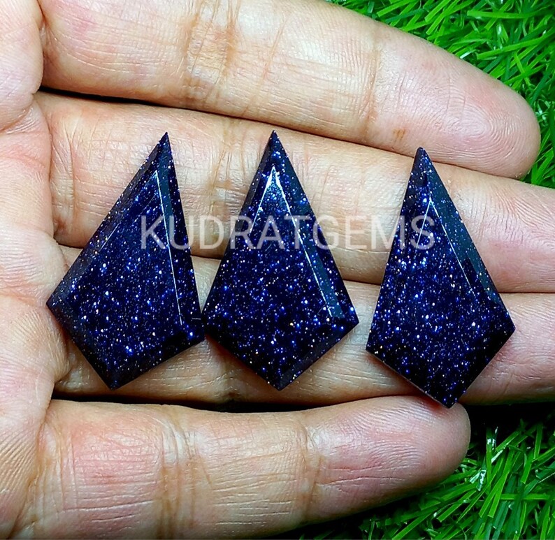 May include: Three kite-shaped blue goldstone cabochons with a deep navy color and sparkling metallic inclusions. Each gemstone features a faceted surface and a pointed top. The text KUDRATGEMS is overlaid across the center of the stones.