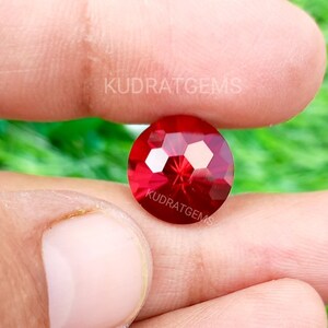 May include: A round, faceted, vibrant red gemstone with a brilliant cut, displaying multiple reflective surfaces. The stone features the text KUDRATGEMS printed twice in white across its surface, indicating the brand name on this loose gemstone.