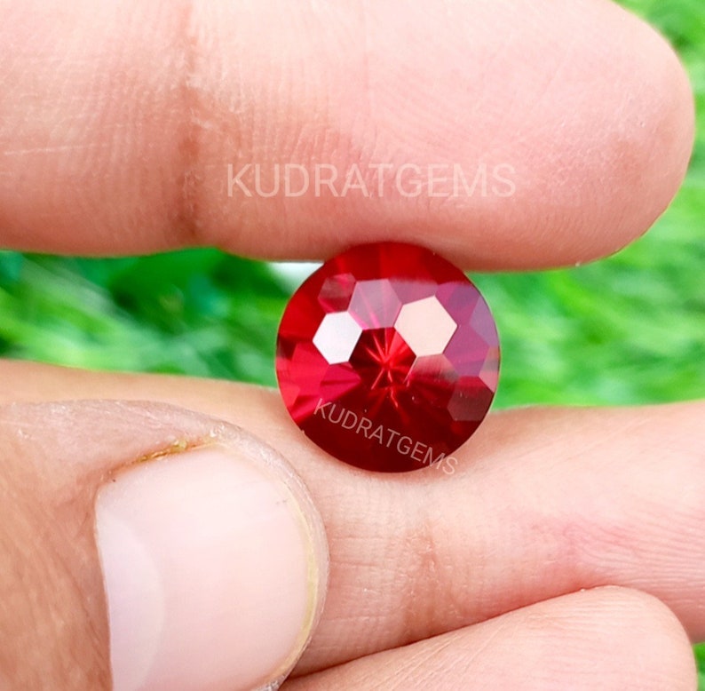 May include: A round, faceted, vibrant red gemstone with a brilliant cut, displaying multiple reflective surfaces. The stone features the text KUDRATGEMS printed twice in white across its surface, indicating the brand name on this loose gemstone.