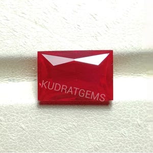Könnte beinhalten: Ein leuchtend roter, rechteckiger Edelstein mit facettierter Oberfläche. Der Edelstein hat ein poliertes Aussehen und trägt den weißen Text "KUDRAT GEMS". Der Edelstein ist vor einem weißen Hintergrund platziert.
