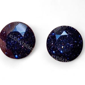 Puede incluir: Dos gemas redondas y facetadas con un aspecto azul oscuro y moteado dorado. Las gemas tienen una superficie pulida y están sobre un fondo blanco. El texto "KUDRAT GEMS" es visible en la superficie de las gemas.