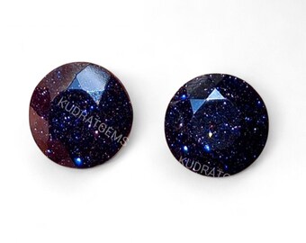 May include: Two round, faceted gemstones with a dark blue and gold speckled appearance. The gemstones have a polished surface and are set against a white background. The text "KUDRAT GEMS" is visible on the surface of the gemstones.
