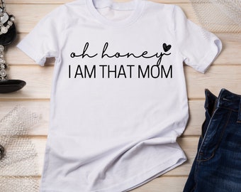 Könnte beinhalten: Weißes T-Shirt mit dem Text "oh honey" und einem kleinen Herzsymbol, gefolgt vom Text "I AM THAT MOM".