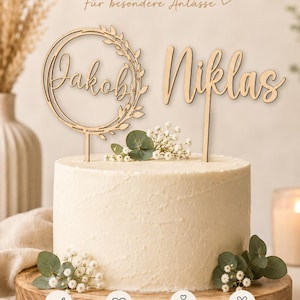 Könnte beinhalten: Ein heller Holzkuchenstecker mit dem Namen Jakob in einem Blattkranz und dem Namen Niklas in Schreibschrift, platziert auf einer weiß glasierten Torte, ergänzt durch Text und Symbole im oberen und unteren Bereich.