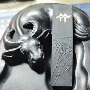 May include: Black ink stick with a carved design of bamboo and the Chinese characters "双溪" (Shuang Xi) in gold. The ink stick is resting on a black stone sculpture of a buffalo.