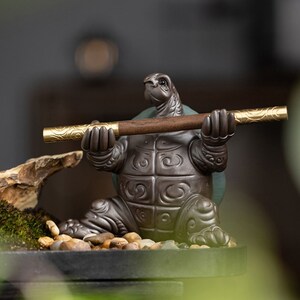 May include: A ceramic turtle figurine holding a wooden and gold incense stick. The turtle is sitting on a small, black base with pebbles and moss around it.