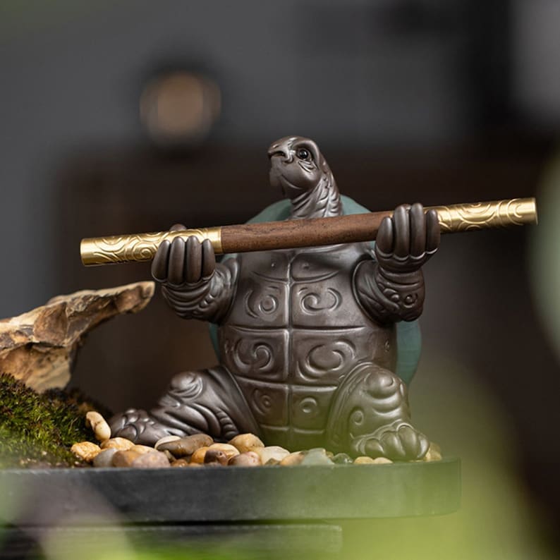 May include: A ceramic turtle figurine holding a wooden and gold incense stick. The turtle is sitting on a small, black base with pebbles and moss around it.