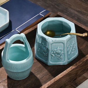 May include: Two light blue ceramic tea ceremony containers. One is a round bowl with a gold spoon inside. The other is a small bucket with a handle.