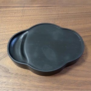 May include: Black ink stone with a wavy, irregular shape. The stone is made of a smooth, dark material and has a slightly textured surface.