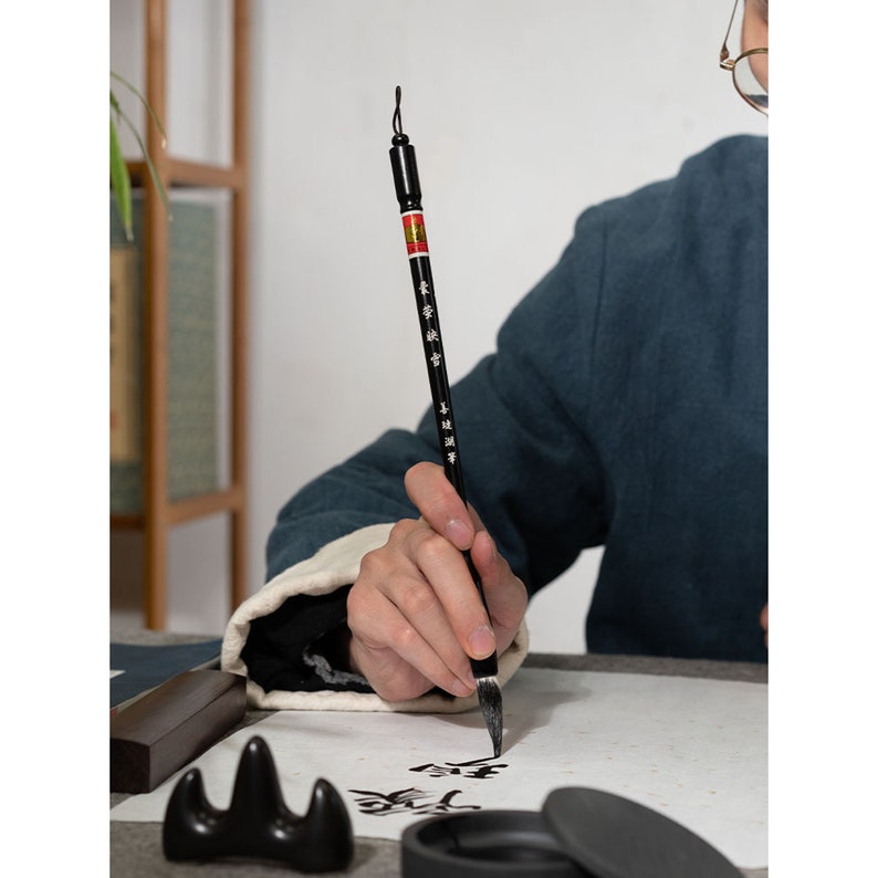 May include: A person is using a black calligraphy brush with a red and black handle to write Chinese characters on white paper. The brush has Chinese characters written on the handle. There is a black inkwell and a black ink stone on the table.