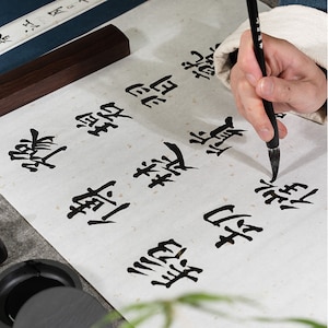 May include: A person writing Chinese calligraphy with a black brush on white paper. The paper has a speckled texture. The person is wearing a light brown shirt. The calligraphy says "樂觀" which means "optimistic".
