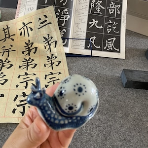 May include: A blue and white porcelain snail figurine with a detailed design. The snail is sitting on a hand.