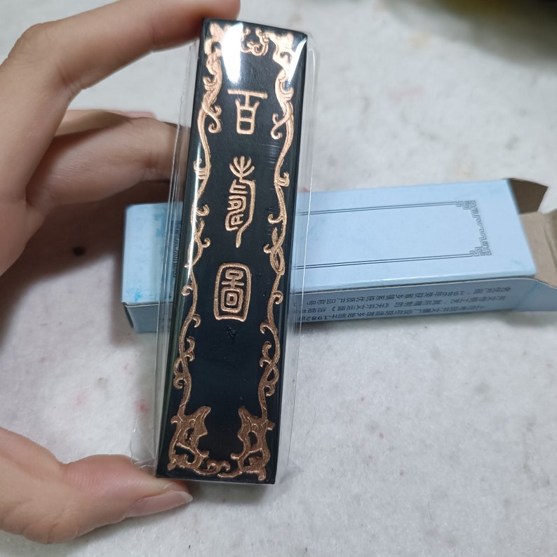 May include: A black ink stick with gold lettering and decorative borders. The ink stick is wrapped in clear plastic and has the Chinese characters "回" and "墨" written on it.