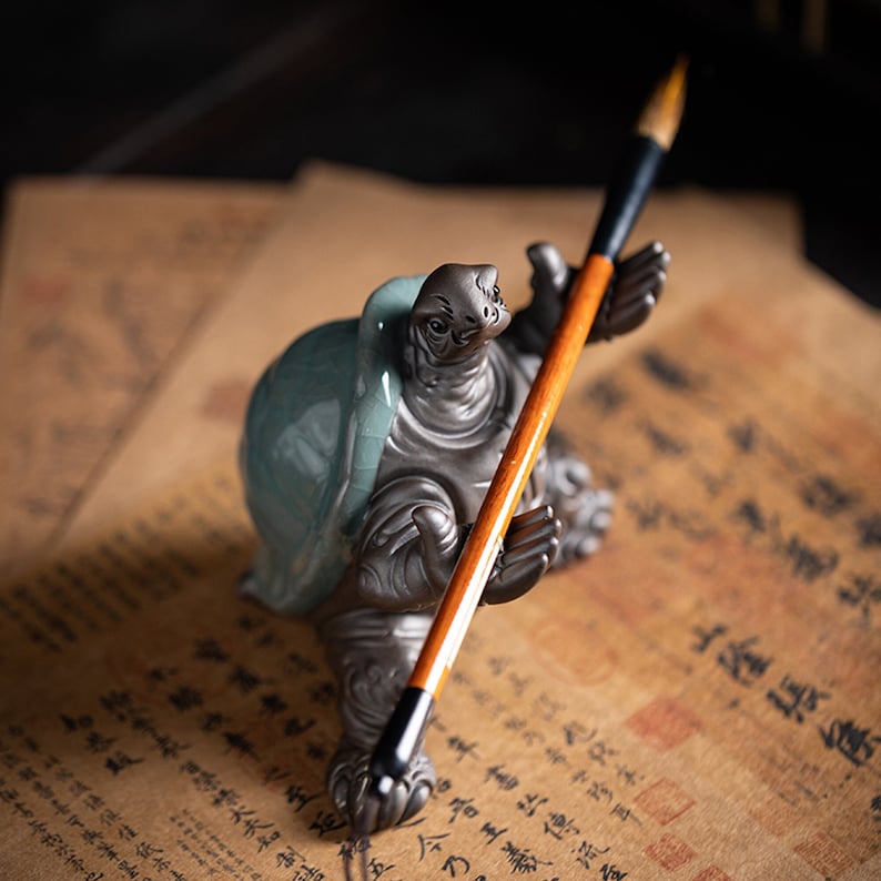 May include: A ceramic turtle figurine holding a brown and black brush. The turtle is painted in shades of gray and blue, and the brush has a long, thin handle.