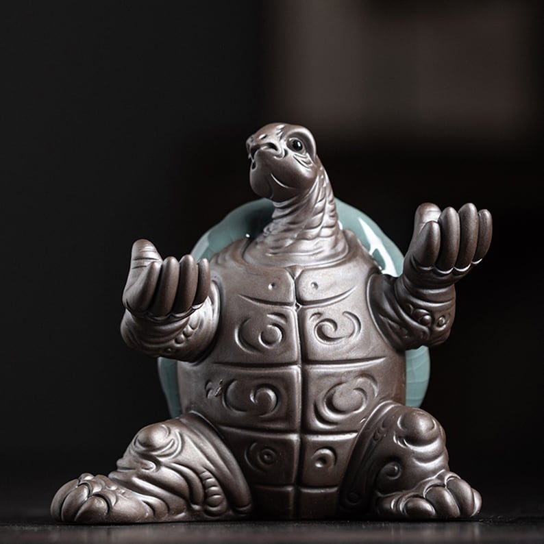 May include: A ceramic turtle figurine in a seated meditation pose with detailed carvings on its shell and limbs. The turtle is brown with a green glaze on its back.