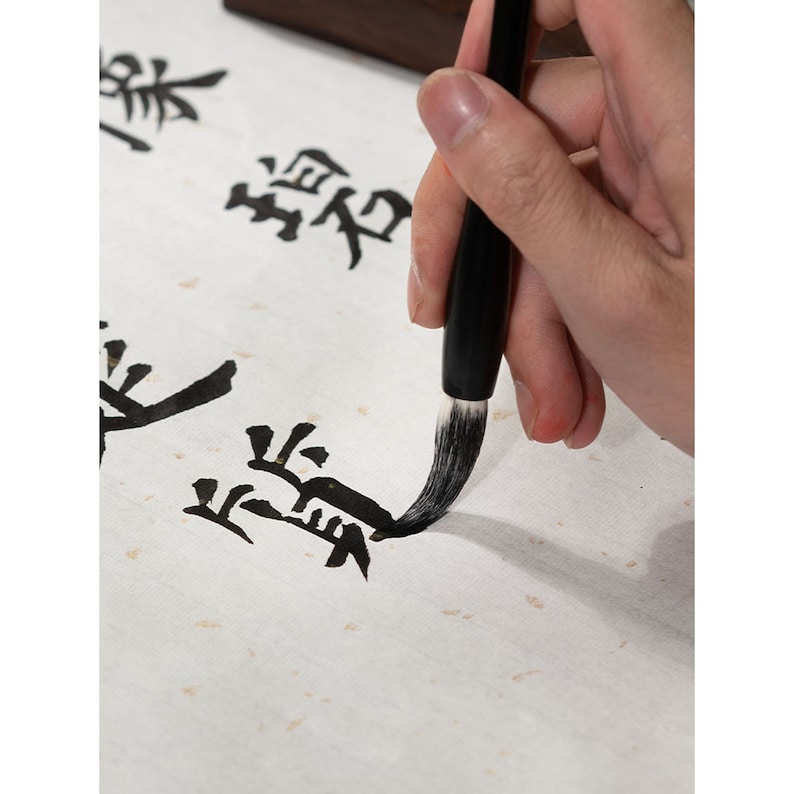 May include: A person's hand holding a black calligraphy brush with white bristles, writing Chinese characters on white paper.