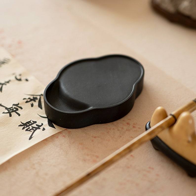 May include: A black inkstone, a traditional Chinese writing tool, sits on a piece of paper with black calligraphy. A wooden brush rests on a stand nearby.