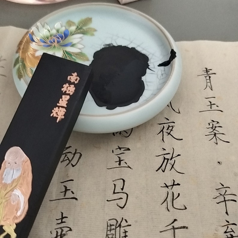 May include: A black ink stick with gold lettering that reads "極品墨寶" in Chinese characters. The ink stick is resting on a white porcelain inkwell with a floral design. The inkwell is filled with black ink. The ink stick and inkwell are on a piece of paper with black calligraphy writing.