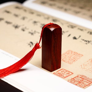 May include: A dark red wooden stamp with a red tassel. The stamp is on a white surface with red Chinese characters.